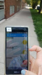 Hand holding a cell phone on a city sidewalk. Air data app shows yellow and red circles indicating moderate levels of particulate matter in the air