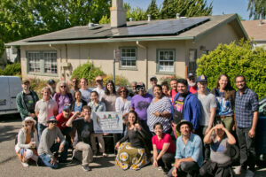 A diverse group of people smiles in front of a home with rooftop solar