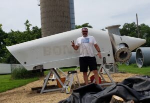 A man in a white t-shirt holds up a peace sign in front of a wind turbine engine on the ground