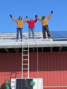 Three men on a rooftop celebrate with their arms up, as they install solar panels. There are some panels installed on the roof behind them, and another on its way up the ladder beneath them.