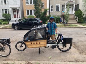 A person stands behind a front-loading cargo bike with a dog inside a rain cover