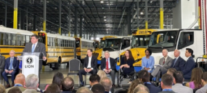 In a large warehouse, a row of electric school buses in the background while a group of people sits listening to Illinois Governor Pritzker speak
