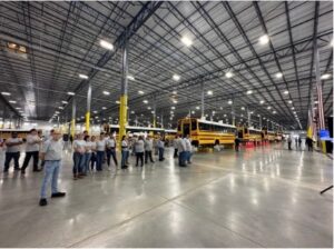 A large sparkly new warehouse has rows of electric school buses in the background and a row of workers standing in the foreground.