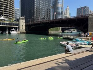 A group of people on green kayaks passes under the State Street bridge of the Chicago River, while onlookers view from the Chicago Riverwalk
