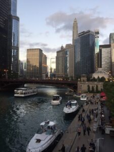 People walk along the Chicago riverwalk, as boats pass by and sun sets in the background