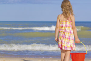 Blonde girl holding a red bucket stands in front of waves at the beach