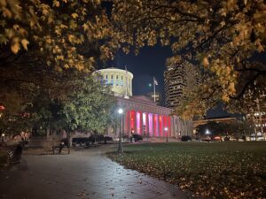 Ohio statehouse in Columbus at night, lit up with red and white lighting amid fall foliage