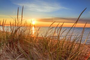 Sunrise paints the blue sky in yellow and orange over sand dunes, grasses, and waves in Lake Huron in Port Austin Michigan