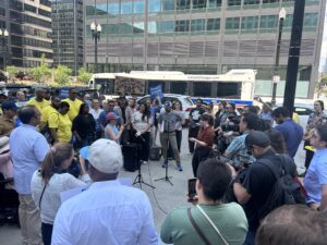 State lawmakers, transit workers, and environmental advocates join everyday riders to advocate for public transit outside Chicago's Union Station, as a CTA bus rolls by in the background.