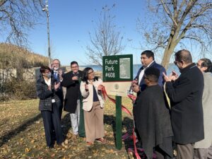 A crowd of people applauds around a green and tan sign that reads "Future Park #608" along Lake Michigan by trees and grass