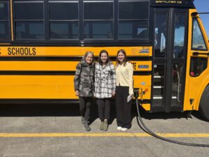 ELPC staff Susan Mudd and Katie Duckworth smile with Spring Intern and Grand Rapids alum Terra Workman in front of an electric school bus plugged in