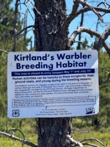 Sign in a tree reads Kirtland's warbler breeding habitat. This area is closed to entry between May 1st and July 15th. Human activities can be harmful to these songbirds, their ground nests, and young during the breeding season.